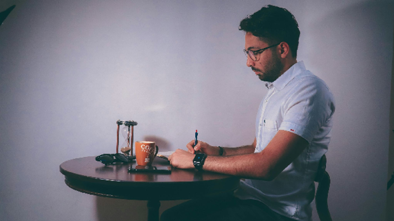 A man wearing glasses writes at a round table next to a coffee mug and an hourglass, focusing on his routine.