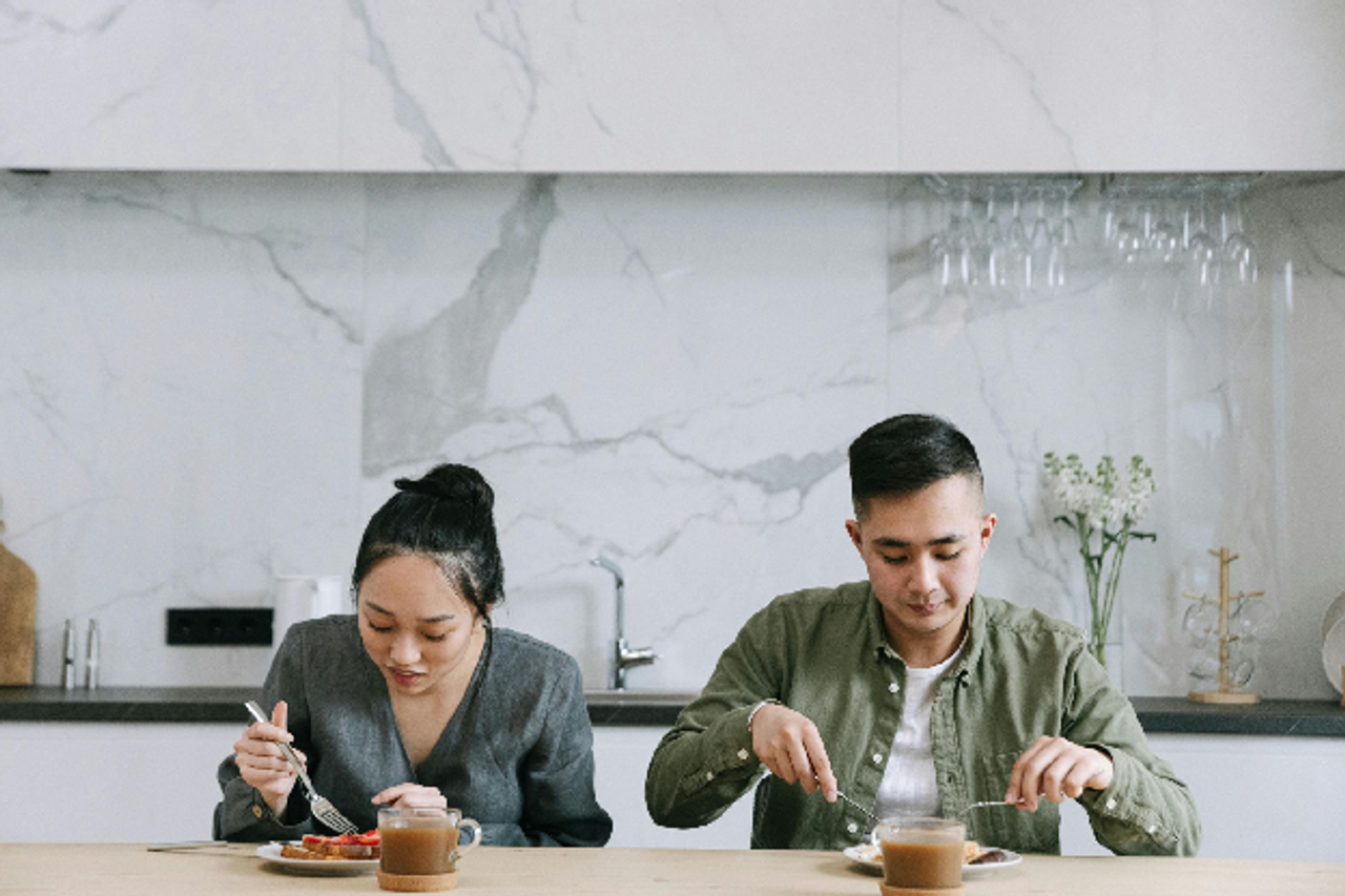 A young man and woman sit side-by-side at a kitchen counter, eating a healthy breakfast in a bright, modern home.